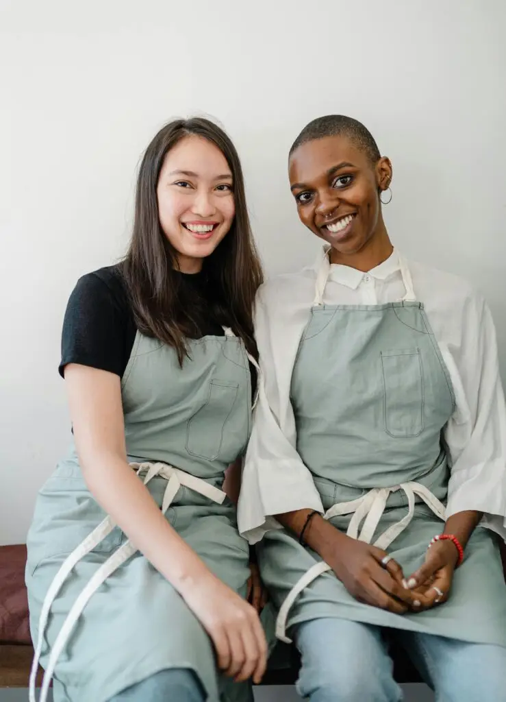 Two diverse women in aprons smiling while sitting indoors, showcasing friendship and happiness.